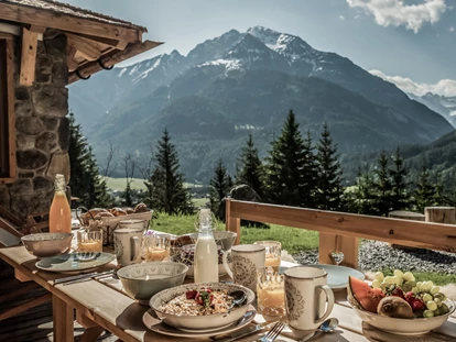 Hüttendorf - Schröcken - Almfrühstück auf der Terrasse im Berg-Chalet. Ein echtes Highlight im Benglerwald Berg Chaletdorf ist das liebevoll zubereitete Almfrühstück, das direkt ans Chalet geliefert wird – mit regionalen Spezialitäten, frischen Zutaten und herrlichem Ausblick auf die Lechtaler Alpen. - Benglerwald Berg Chaletdorf